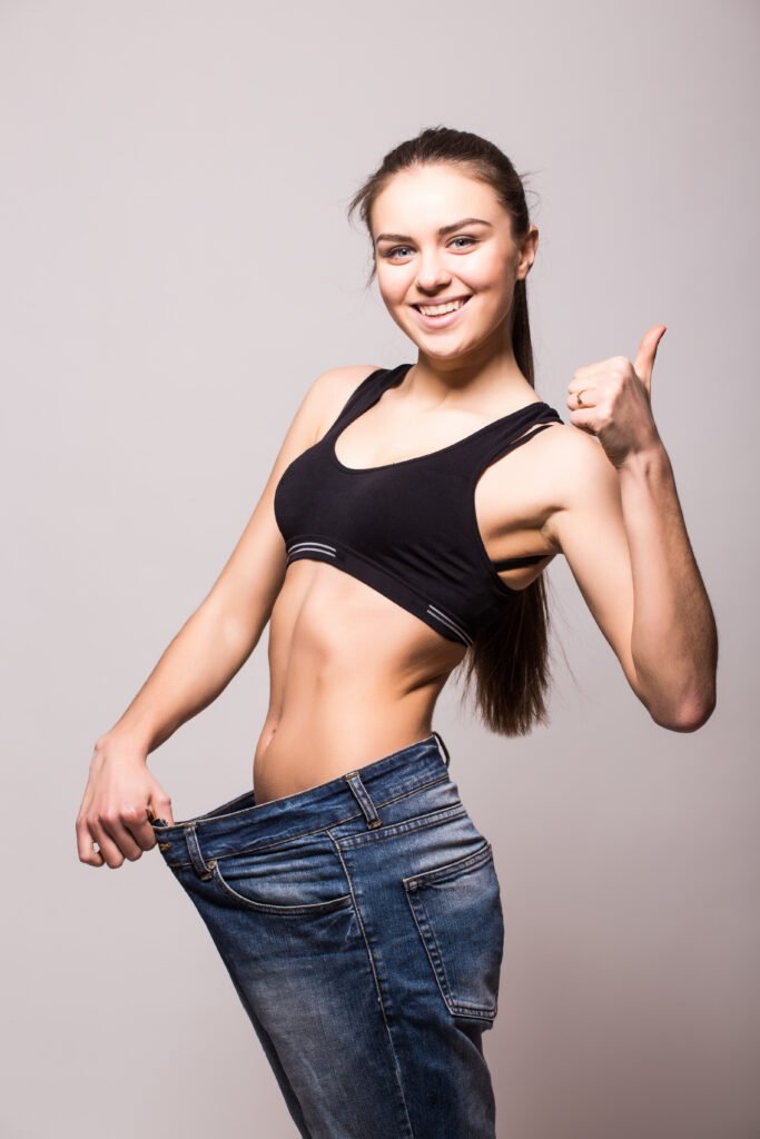 Funny woman shows her weight loss by wearing an old jeans, isolated on white background